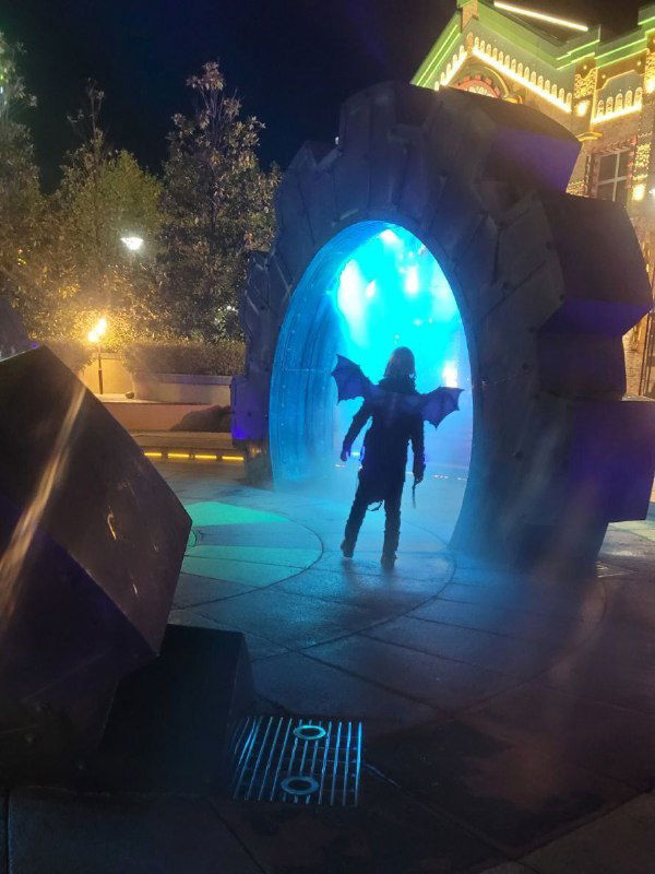 Силуэт человека у подсветки и арки в парковой зоне — атмосферный ночной кадр с иллюминацией и туманом на хэллоуинской локации.