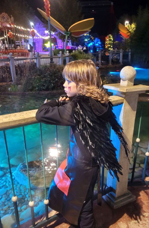 Ребёнок в тёмном плаще с перьями опирается на перила у водоёма, на заднем плане огни и декор Сочи‑парка в ночь Хэллоуина.