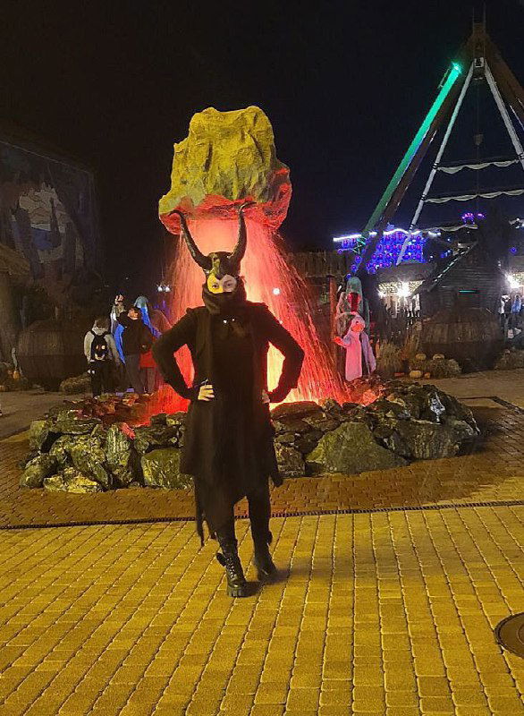 Человек в чёрном костюме с яркой жёлтой маской на фоне сценического декора и инсталляций в парке — ночной хэллоуинский сценический кадр.