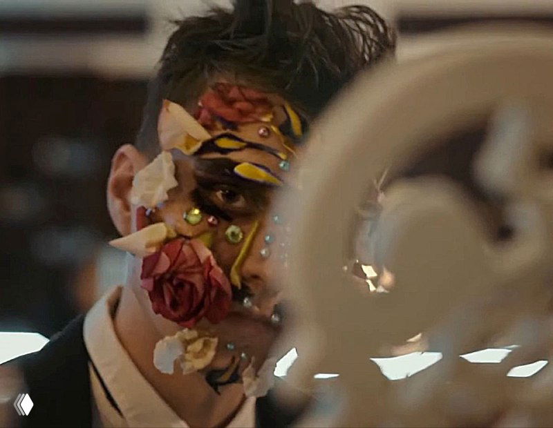 Мужчина в смокинге с украшениями из лепестков и страз на лице, крупный план декоративного макияжа и цветочных элементов в клипе