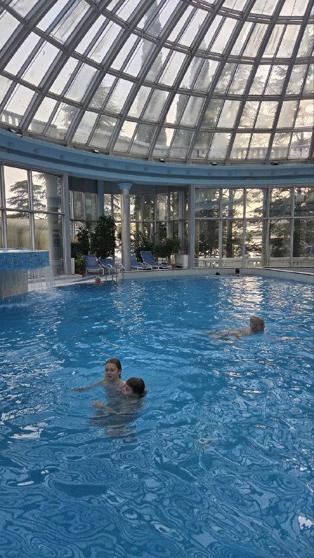 Люди плавают в бассейне под стеклянным куполом: взрослый с ребёнком у борта, отражения купола в воде и спокойная атмосфера.