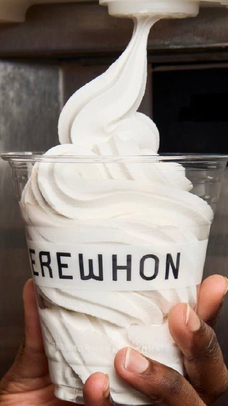 Крупный план стаканчика мягкого мороженого с надписью 'EREWHON' в руке — контраст белой текстуры и тёмного фона, бытовая деталь городского ландшафта.