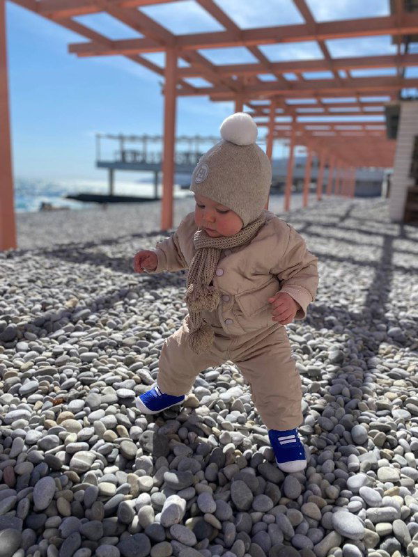 Малыш в тёплом комбинезоне делает первые шаги по гальке у перголы на пляже; видны море и солнечное небо, семейная прогулка.