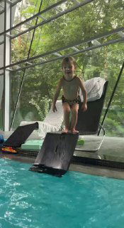 Ребёнок на бортике бассейна внутри комплекса на фоне больших окон с видом на лес, готовится к прыжку или игре в воде.