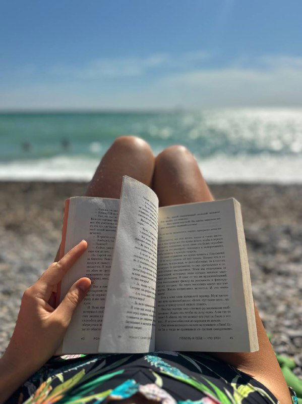 Крупный план раскрытой книги на коленях у человека на пляже, галька под ногами и море на заднем плане — спокойный момент чтения на отдыхе.