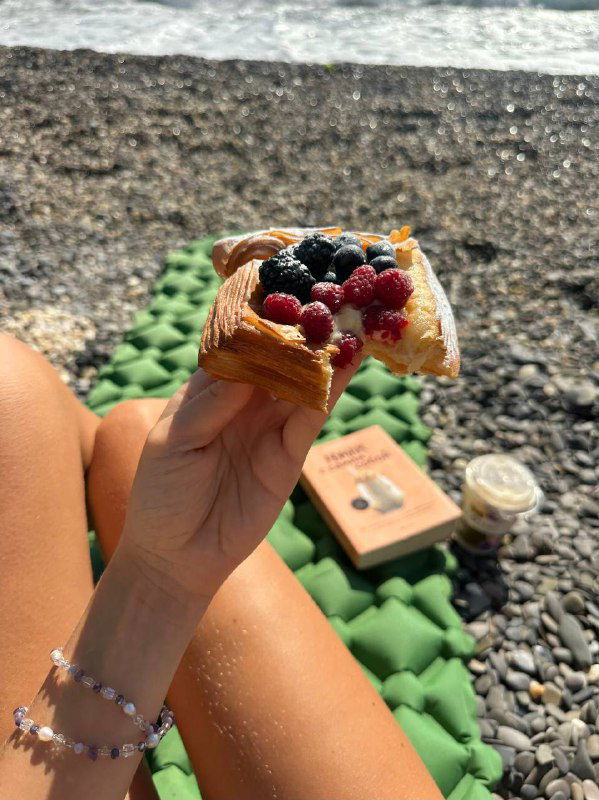 Рука держит кусочек выпечки с ягодами на фоне пляжного коврика и книги; летнее угощение во время отдыха у моря, кадр с деталями.