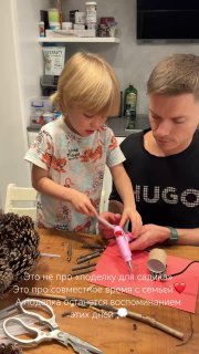 Ребёнок и отец за столом делают поделку; ребёнок использует клеевой пистолет, вокруг — материалы для творчества и шишки.