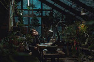 Кадр из фильма: мастерская или оранжерея с деталями декора, герой сидит за столом в задумчивой позе — тёмная атмосферная сцена.