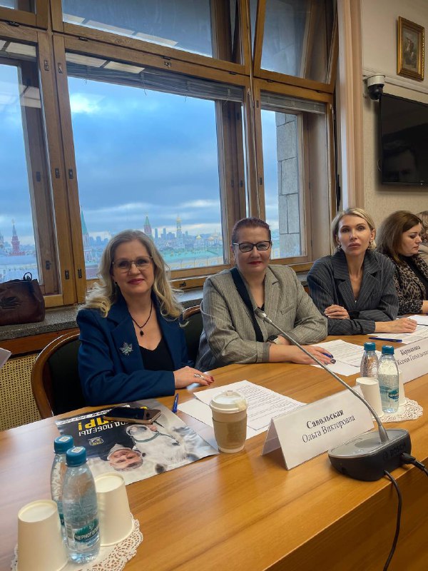 Автор и коллеги сидят за столом в переговорной с видом на город через большое окно; на столе документы, бутылки с водой и чашка кофе.