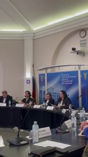 Панельная дискуссия или круглый стол: несколько спикеров за столом в конференц-зале, микрофоны и бутылки воды, деловая обстановка.