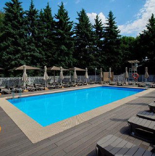 Бассейн на деревянной палубе с зонтиками и шезлонгами, окружён зелёными деревьями; дневная летняя атмосфера отдыха в городском парке Москвы.