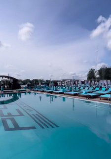 Большой открытый бассейн с бирюзовой водой, многочисленные шезлонги и белые зонты; спокойная пляжная зона у реки для отдыха и фото.