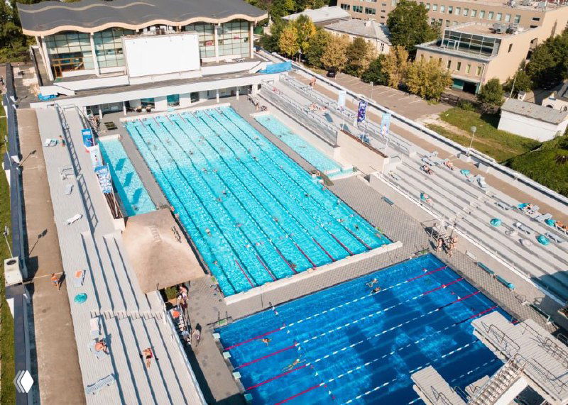 Спортивный бассейн с дорожками для плавания, трибунами и открытой территорией — вид сверху дрона на комплекс бассейнов и прилегающие зоны.