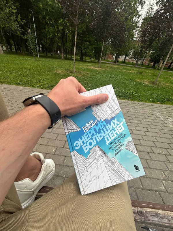 Книга «Энергия больших денег» Райана Серханта в руках автора на скамейке в парке, видна обложка с графикой и городской пейзаж.