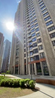 Фасад многоэтажной новостройки в Москве на солнечном дне; видны входная группа, окна этажей и часть благоустроенной придомовой территории.