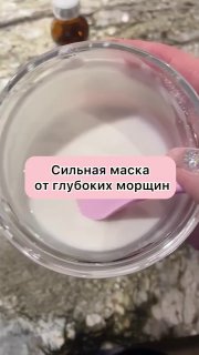 Вид сверху: прозрачная чашка с белой, плотной смесью маски; внутри розовая лопатка, на заднем плане размытая бутылочка.