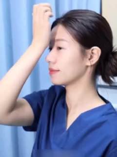 Женщина в синем халате демонстрирует гимнастику для лица в боковом профиле; рука у лба, спокойный фон штор, мягкое освещение.