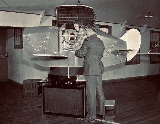 Ранний Link Trainer на подвижной платформе: вид сбоку с открытой кабиной и инструктором рядом, историческая чёрно‑белая фотография учений.