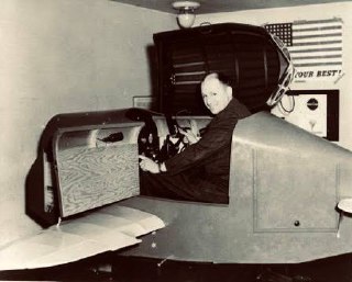 Исторический кадр с Link Trainer: мужчина в кабине тренажёра демонстрирует органы управления, на заднем плане элементы учебного зала и флаг.