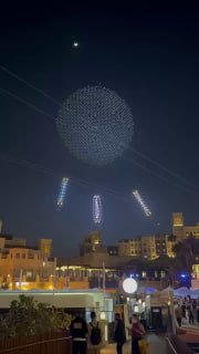 Ночное небо Дубая: сотни светящихся дронов формируют крупный круг над набережной, видны огни и силуэты зрителей и зданий.
