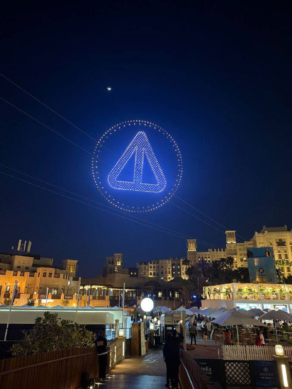 Треугольный логотип, собранный из множества дронов в ночном небе Дубая; вид с улицы на зрителей и подсветку набережной.