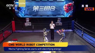 Женский комментатор или ведущая стоит в ринге между двумя маленькими гуманоидными бойцами; по бокам сидят зрители, на заднике показан экран с эмблемой.