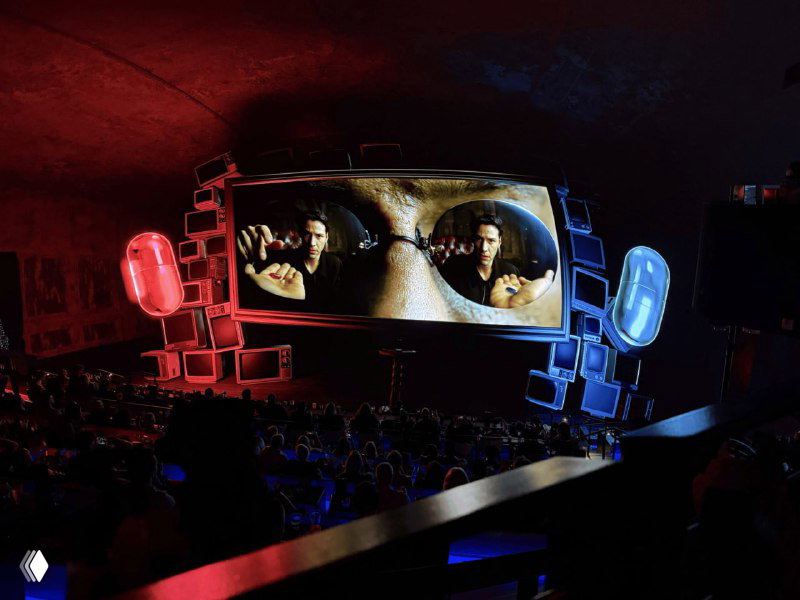 Широкий кадр зала с большим экраном, на котором видны очки и отражения — стилизованная сцена из «Матрицы», окружённая цветными подсветками.