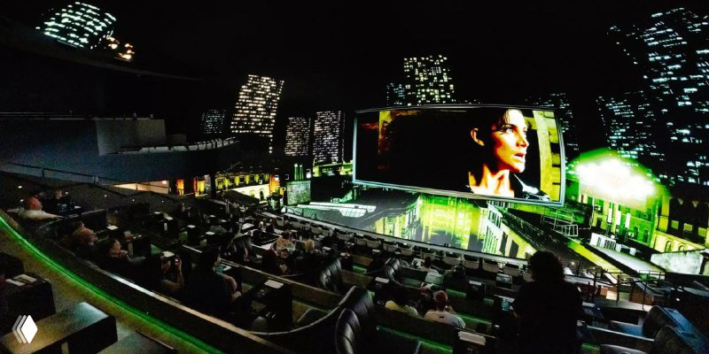 Зрители в ряду наблюдают за панорамной проекцией; на экране крупный план героини, окружающие проекции охватывают всё пространство зала.