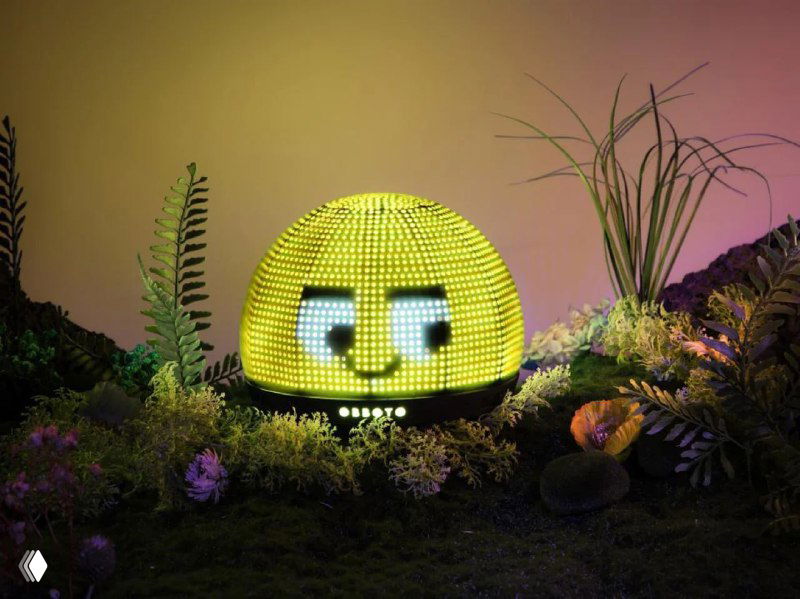 Крупный план круглой светящейся мини‑сферы OBBOTO на фоне декора: на дисплее пиксель‑арт в виде улыбающегося лица, яркие зелёно‑жёлтые пиксели.