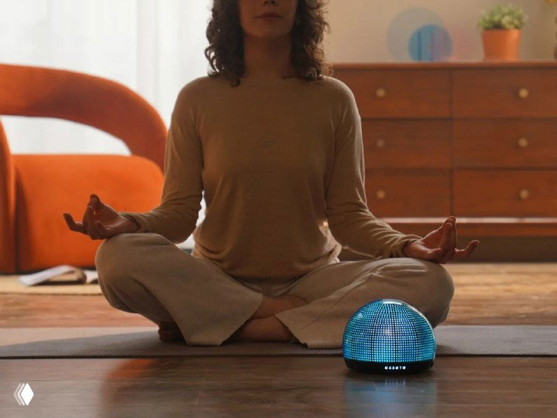 Человек медитирует на коврике в гостиной, перед ним на полу стоит мини‑сфера OBBOTO с мягкой голубой подсветкой, создающая спокойную атмосферу для практики.