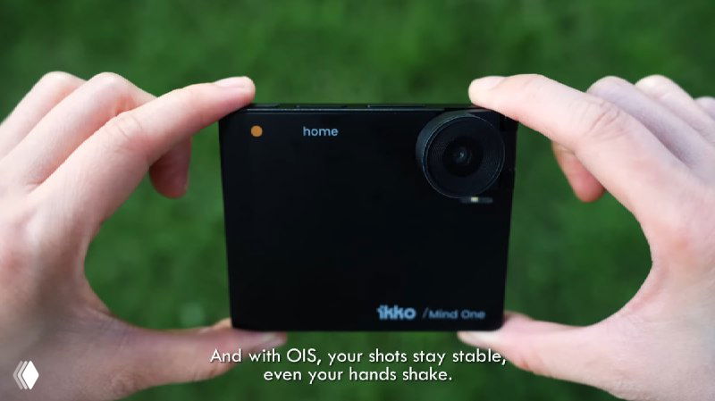 Чёрный iKKO Mind One в руках крупным планом; показана лицевая часть с объективом и маркировка «Mind One» на корпусе в естественной обстановке на зелёном фоне.