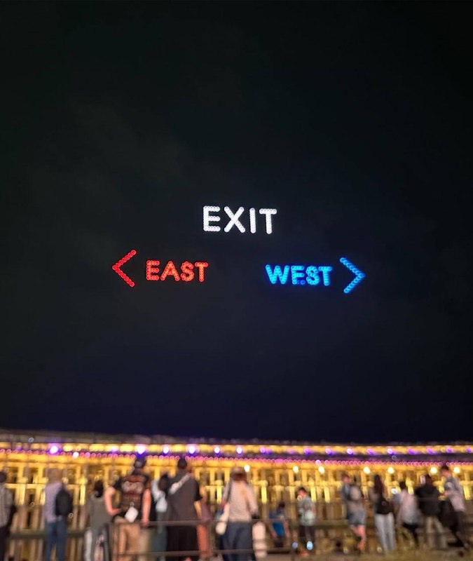 Крупный план ночного неба с надписью «EXIT» и красной/синей стрелкой по бокам; внизу видны зрители и арка стадиона под подсветкой.