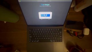 Ночной ракурс сверху: подсветка клавиатуры MacBook, на экране приложение TrackWeight; видны аксессуары рядом, демонстрация работы трекпада как весов.