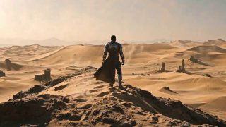 Одинокая фигура мужчины, идущего по дюнам в пустыне; кинематографический пейзаж с подчёркнутой масштабностью и ощущением пути.