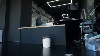 На фото в офисном пространстве маленькое автоматическое мусорное ведро на полу на фоне рабочего стола и человека за компьютером — тест робота в реальной обстановке.