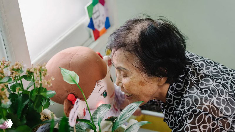 Пожилая женщина целует умную куклу у подоконника; кадр подчеркивает эмоциональную близость и домашний характер использования Hyodol.