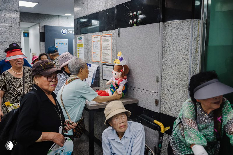 Группа пожилых людей в общественном помещении с куклами Hyodol; сцена очереди и распространения устройства среди пенсионеров.