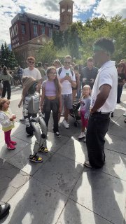 Rizzbot среди прохожих в городском сквере: гуманоид в шляпе окружён людьми разных возрастов, видно реакцию публики и здания.