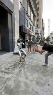 Rizzbot на тротуаре исполняет жесты и позирует прохожим; люди фотографируют робота на телефоны на фоне городских зданий.