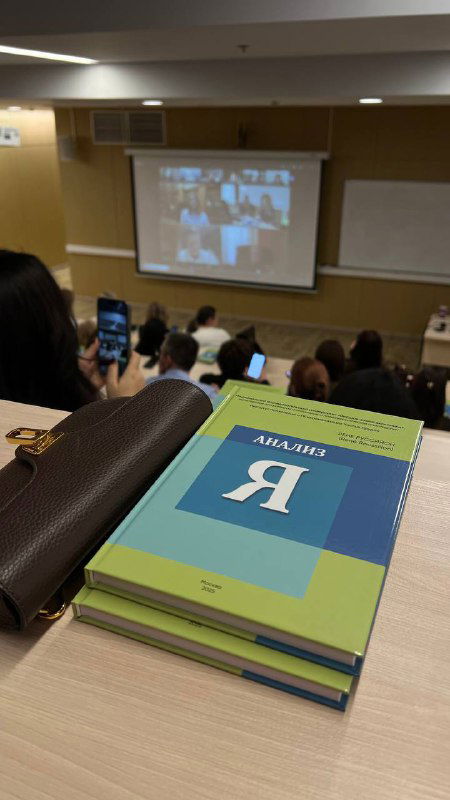 Фото стопки книг «Анализ Я» на столе в лекционной аудитории, на переднем плане обложка переведённой книги и фон с участниками и проекцией трансляции.
