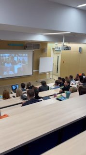 Широкий план лекционной аудитории с участниками презентации, видна проекция докладчика на экране и живая аудитория, слушающая онлайн-включение.