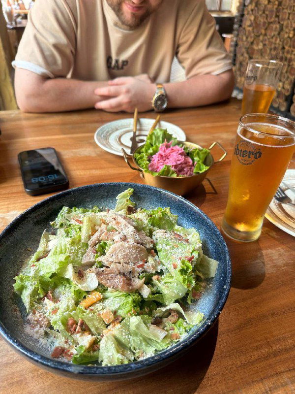 Обед на веранде у моря: большая тарелка салата, миска с закуской и бокал пива на деревянном столе — лёгкий ресторанный перекус в путешествии.