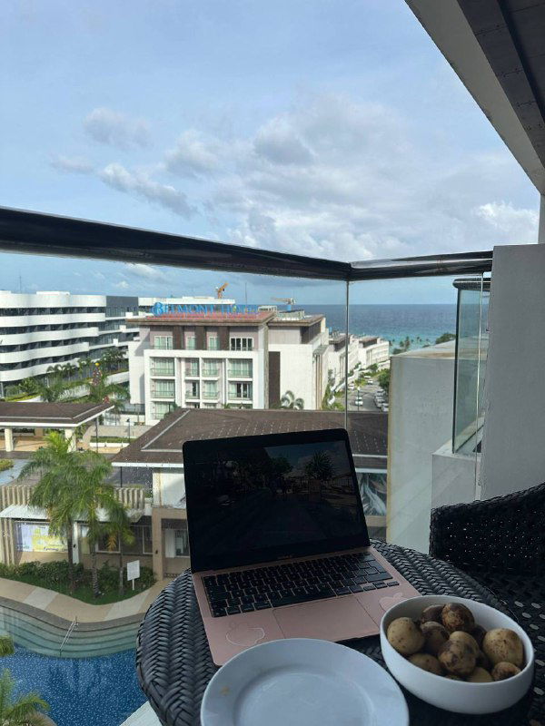 Балконное рабочее место: ноутбук и миска с фруктами на столе, вид на отели и море — сочетание работы и отдыха вдали от дома.
