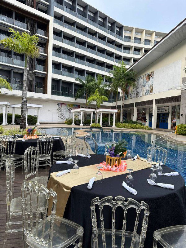 Площадка у бассейна большого курортного отеля с накрытым столом и декором для мероприятия, вокруг пальмы и многоквартирный корпус отеля.