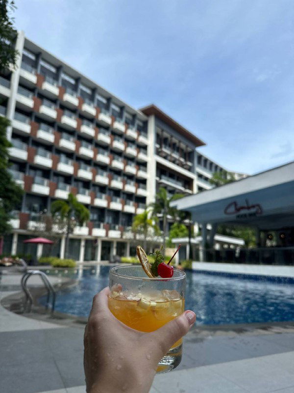 Крупный план коктейля на фоне бассейна и фасада курортного отеля; кадр подчёркивает курортную атмосферу и расслабление у воды.