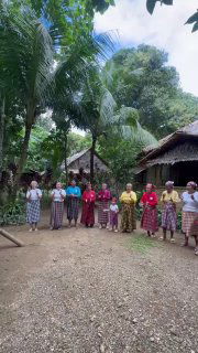 Несколько местных жителей стоят в ряд перед домиками среди тропической растительности: женские и мужские фигуры в традиционной одежде на ферме