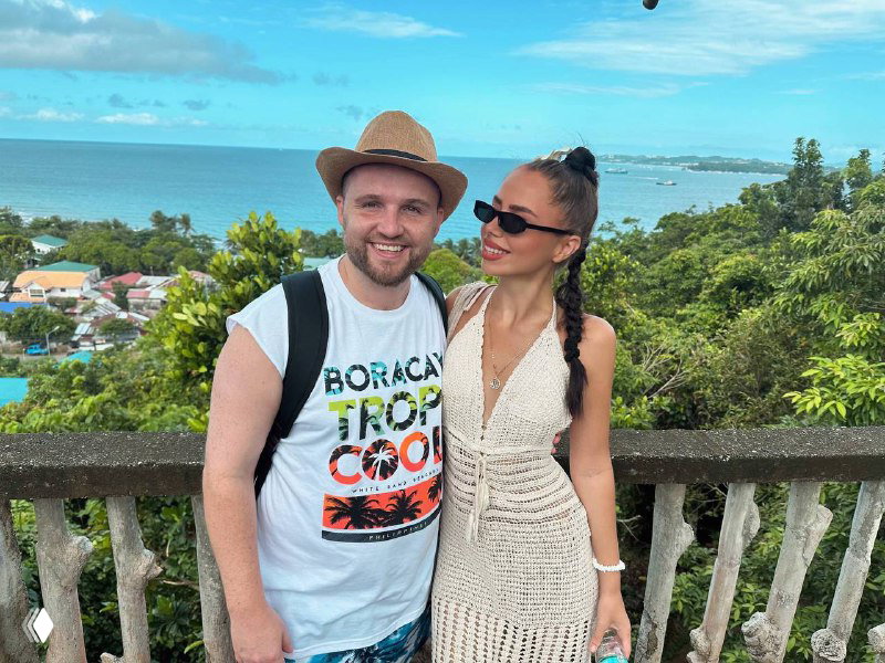 Пара на смотровой площадке с видом на море и прибрежный поселок: автор в лёгком летнем наряде и спутник на фоне тропического пейзажа