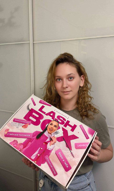 Ученица позирует с закрытым лэш‑боксом розового дизайна с надписью LASH BOSS, коробка занимает центральное место в кадре студии.