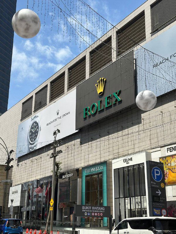 Фасад большого торгового центра в Куала‑Лумпуре с вывеской Rolex и витринами люксовых брендов под дневным небом и декоративными шарами.