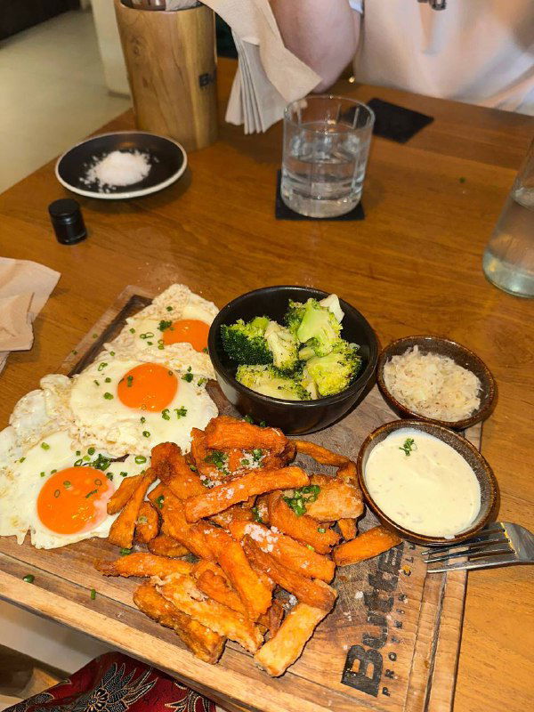 Тарелка с завтраком: яйца, батат-фри, брокколи и соусы на деревянной доске в уютном кафе — пример местной кухни в путешествии.